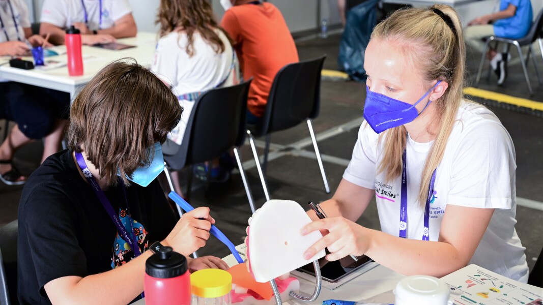 On the picture you see two women, one athlete is brushing teeth of a model set of teeth with the help of a big toothbrush.