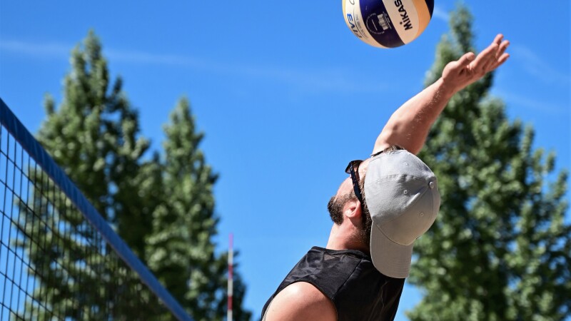 The photo shows a player in action at a beach volleyball competition. He is about to execute an attacking shot. He is wearing a cap and sunglasses.
