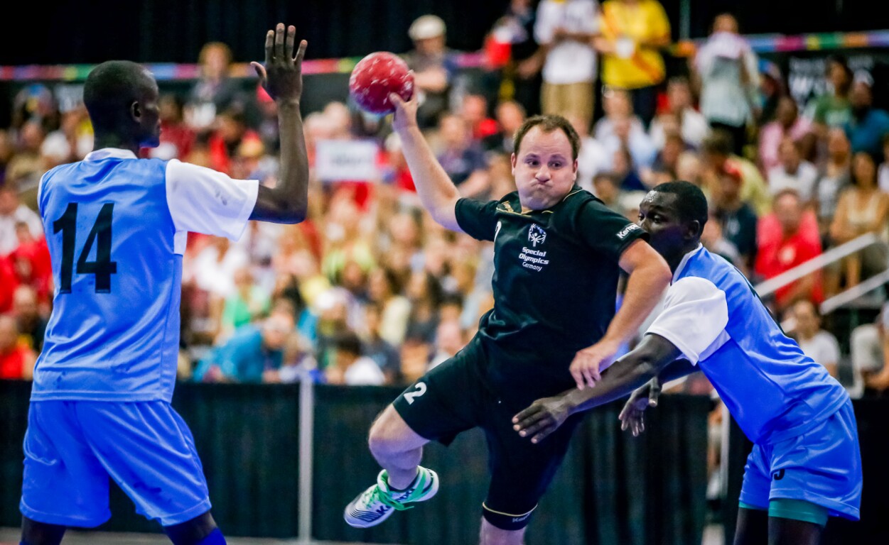 Das Foto ist während eines Special Olympics Handballspiels aufgenommen. Im Vordergrund ist ein Athlet im Sprungwurf und zwei gegnerische Spieler versuchen ihn zu verteidigen. Im Hintergrund sind volle Zuschauerränge.