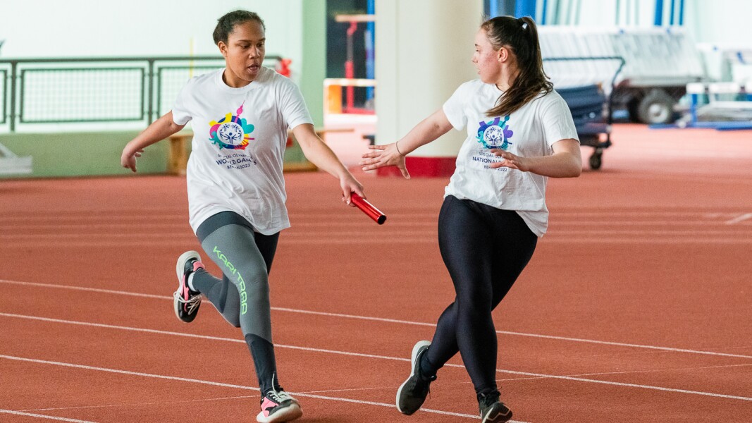 Auf dem Foto sind zwei Athletinnen zu sehen, die im Rahmen der Pressekonferenz eine Sportdemonstration machen. In dem Moment des Bildes übergibt eine Athletin gerade den Staffelstab.