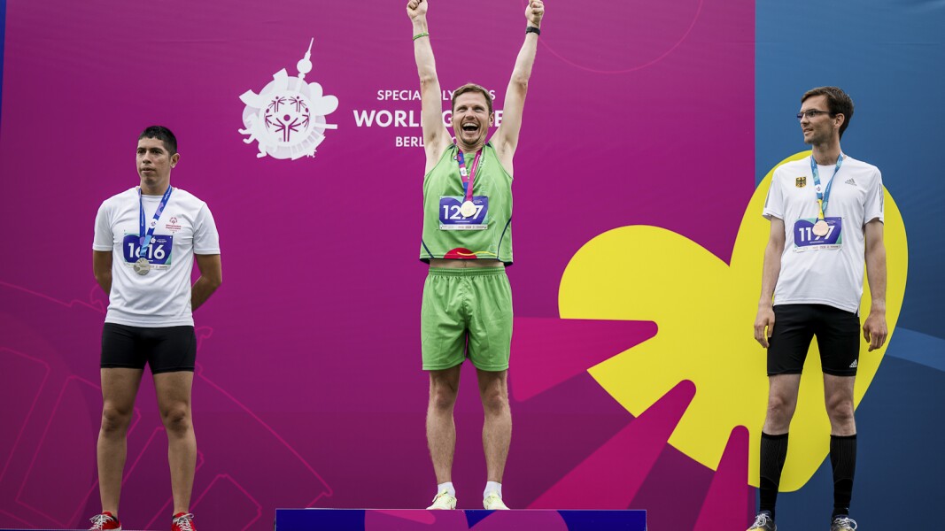 Auf dem Foto ist die Siegerehrung der Männer im 5000m-Lauf. Der Gewinner streckt die Arme in die Höhe.