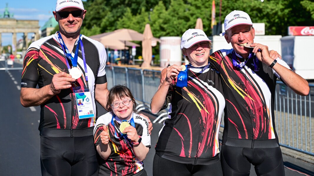 Auf dem Foto sind vier Radsportler*innen zu sehen, die nach ihrem Rennen und der Siegerehrung mit ihren Medaillen posen und glücklich in die Kamera lächeln.