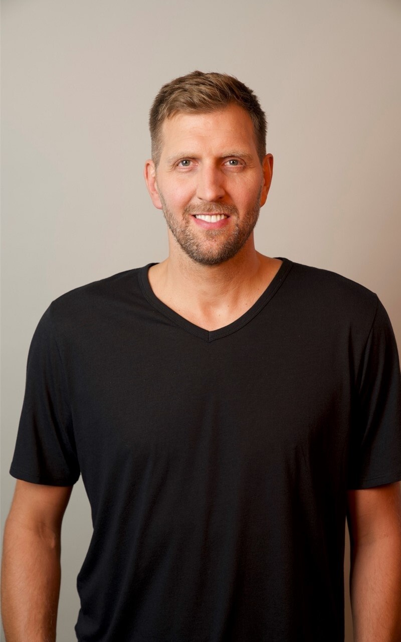 The photo shows the former German basketball player Dirk Nowitzki. He wears a plain black T-shirt and smiles friendly into the camera. 