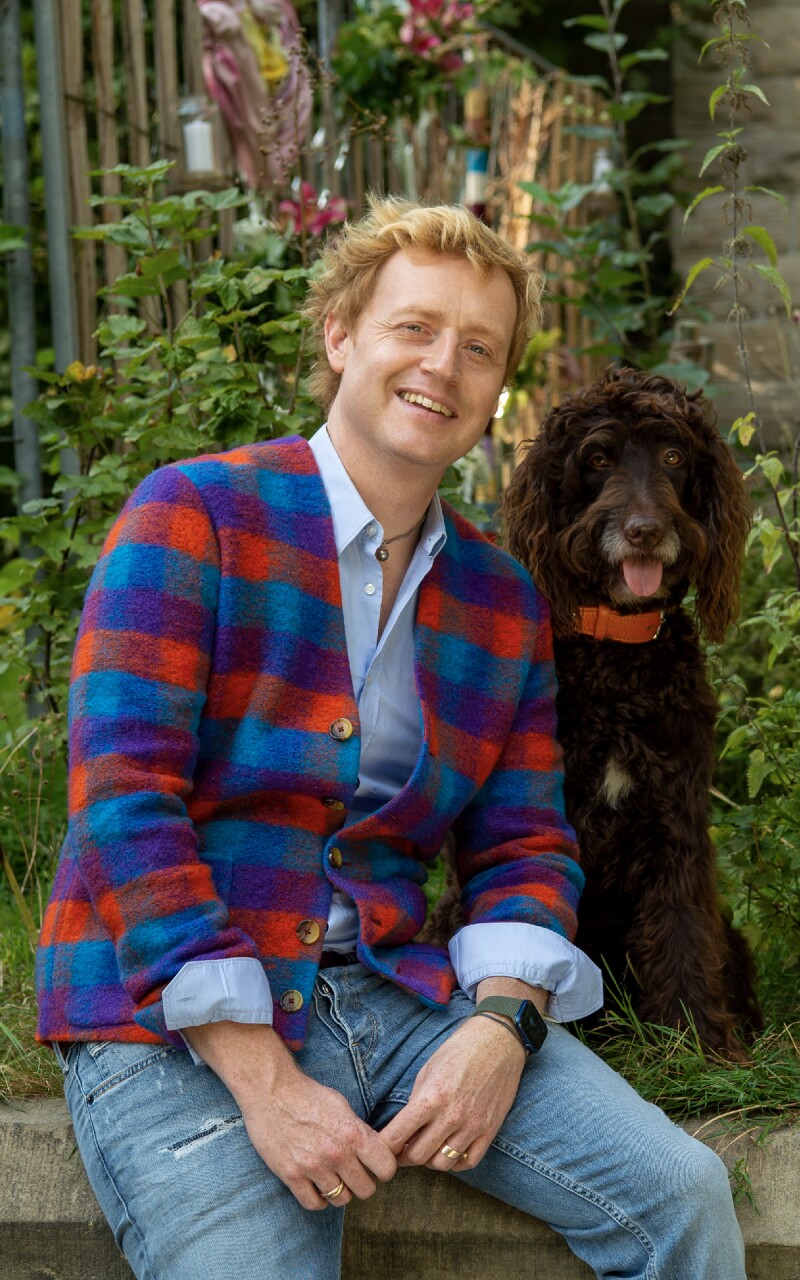 Das Foto zeigt den Floristen Björn Kroner mit seinem Hund Rummel. Kroner trägt ein buntes Sacko und lächelt in die Kamera.