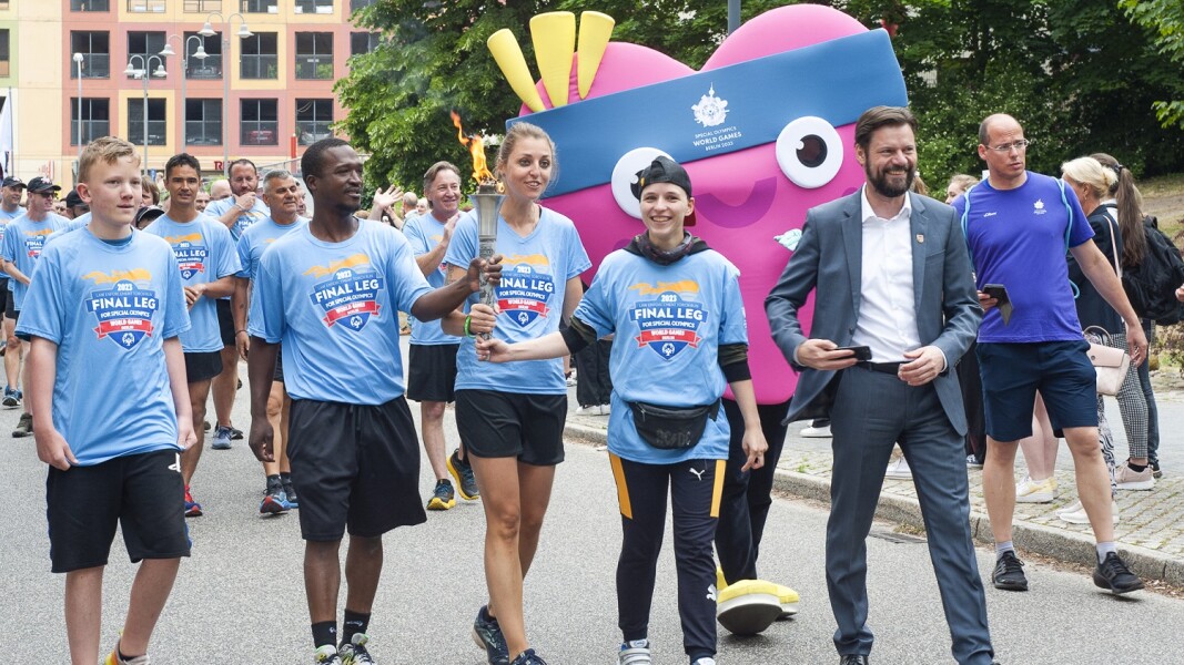 The torch is carried by four people, who are accompanied by the mascot, a local representative wearing a gray suit, and other people in the background.