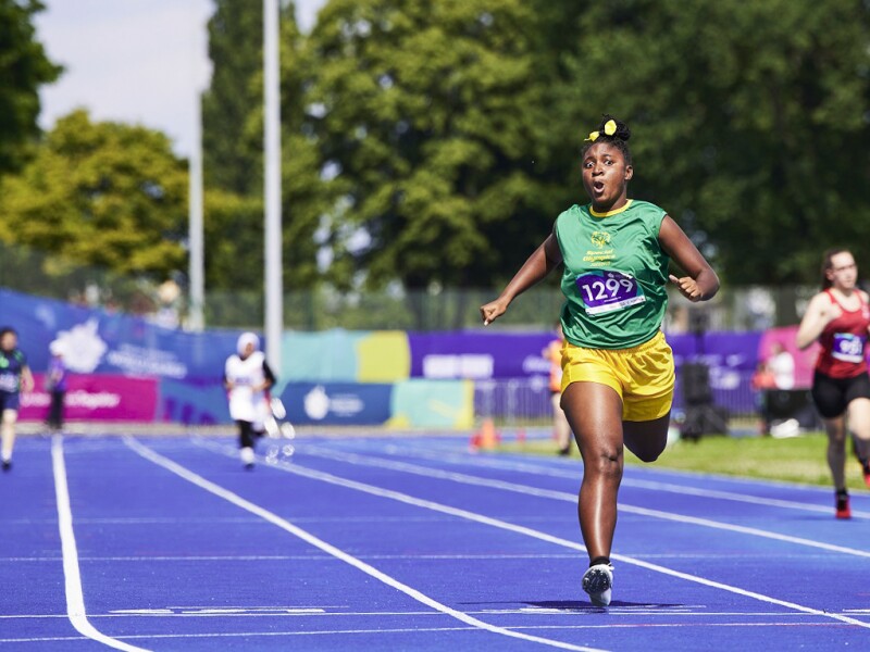 Four athletes running on the blue track while the Jamaican athlete Sheneel Williams in the yellow shorts and green jersey is approaching the finishing line.