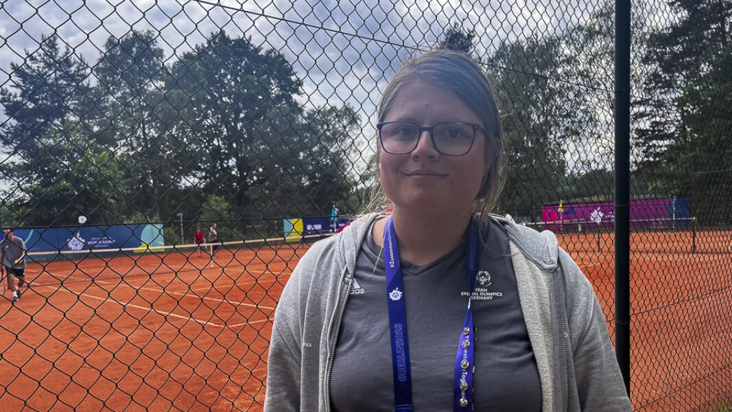 On the photo you can see a tennis player. She is standing in front of the tennis court and is happy.