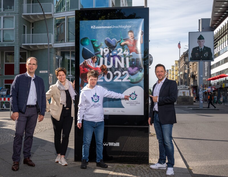 The photo shows an out of home advert poster with our event campaign for Berlin 2022. LOC employee and activist Arthur Hackenthal, CMO Albert Tuemann and two Wall GmbH employees are standing in front of it. The photo was taken directly at the famous Checkpoint Charlie Berlin. 