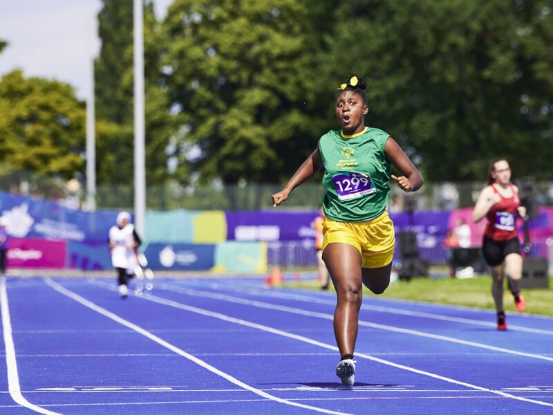 Das Foto zeigt die Tartanbahn bei der Leichtathletik, auf der gerade ein Rennen stattfindet. An der Spitze ist eine Athletin aus Jamaica.