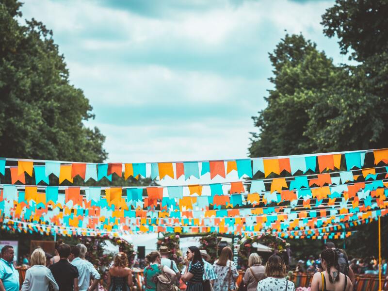 The photo shows a festival scene. Many colorful pennant strings are stretched over the people between two rows of trees.
