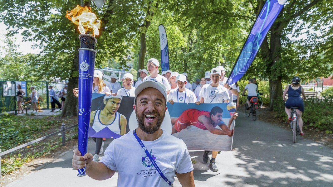 Das Bild zeigt eine Gruppe von Personen beim Fackellauf. Im Vordergrund läuft Paralympics-Athlet Mathias Mester. Er hält eine Fackel in der Hand und lächelt. Dahinter laufen viele Personen die ein Banner und Flaggen tragen.