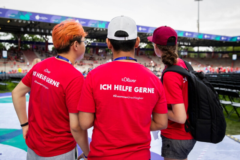 In the photo, three volunteers can be seen from behind during the Opening Ceremony of the National Games. Their T-shirts say "Ich helfe gerne" which translates to "I am happy to help".