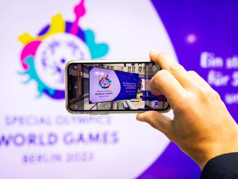 Auf dem Foto, bei dem ein Handy zu sehen ist, welches ein Foto von einer Leihnwand mit der Aufschrift: " Ein starkes Team für Sichtbarkeit und Teilhabe" macht.