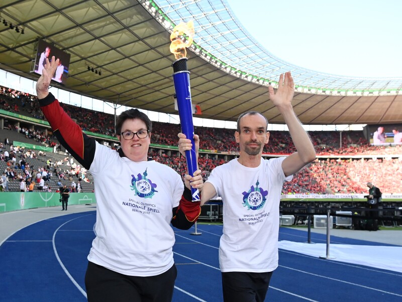 Robert Herberg, Gesicht der Special Olympics Nationalen Spiele Berlin 2022, und Athletin Juliana Rößler halten gemeinsam die Fackel von Special Olympics. Sie laufen auf der blauen Bahn im Berliner Olympiastadion.