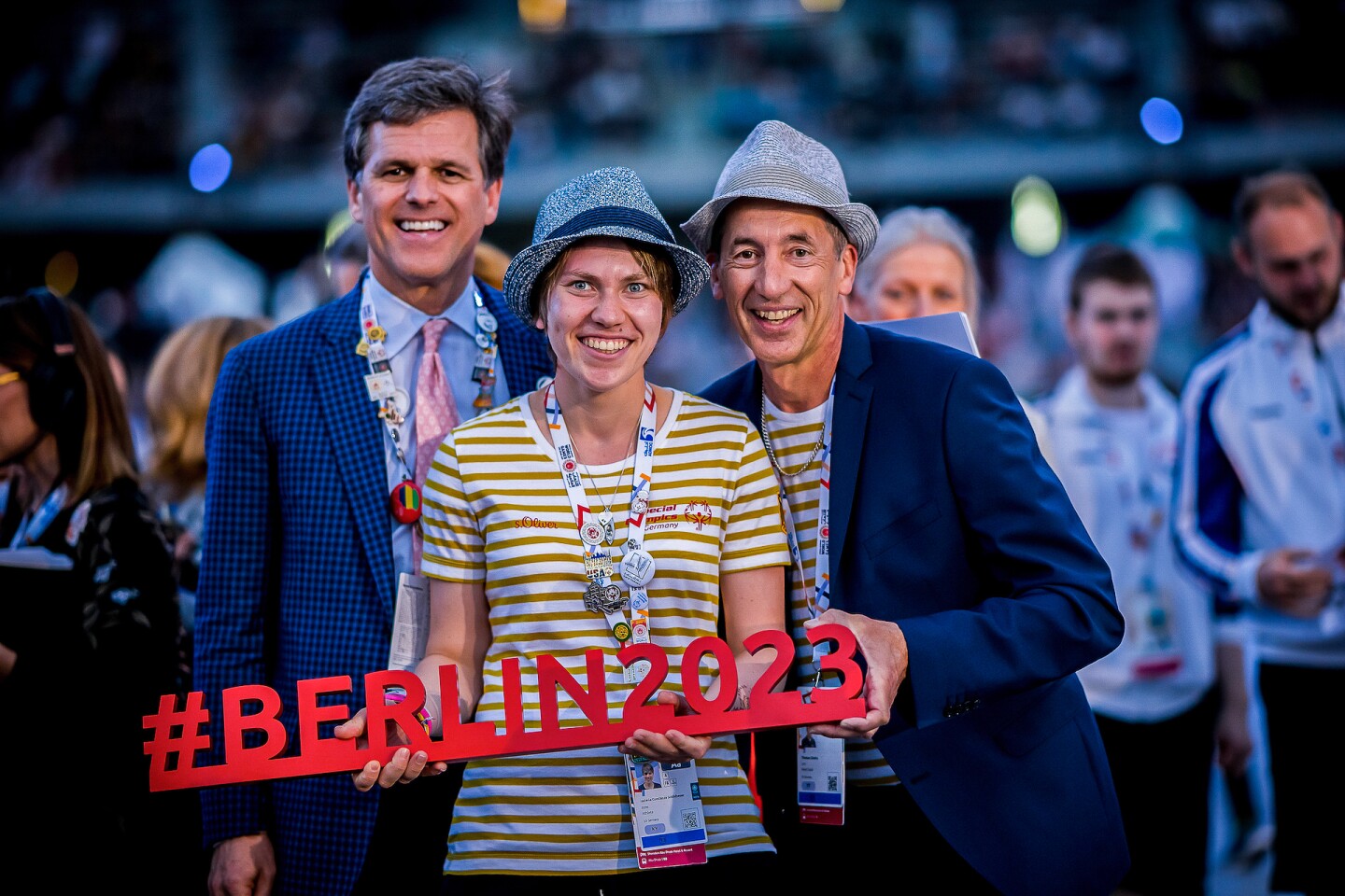 Auf dem Bild sieht man Timothy Shriver zusammen mit Athletin Isabelle Schildheuer und ihrem Trainer Thomas Gindra. Sie halten ein Berlin 2023 Schild in ihren Händen.