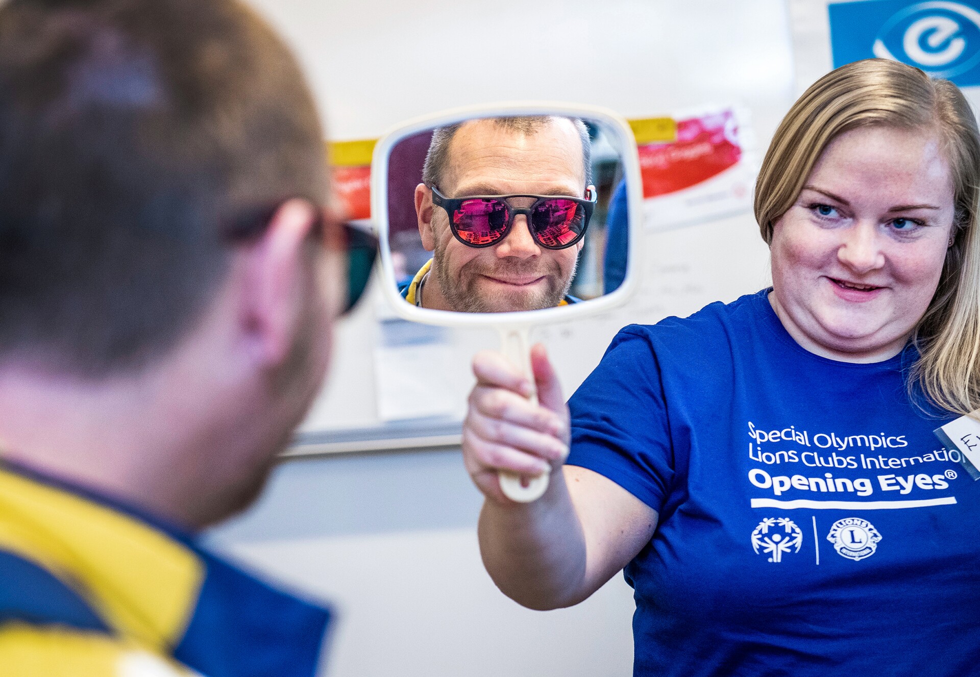 The photo shows a man and a woman during an "Opening Eyes" examination. The woman holds up a mirrow and the man sees himself with sunglasses on while smiling.
