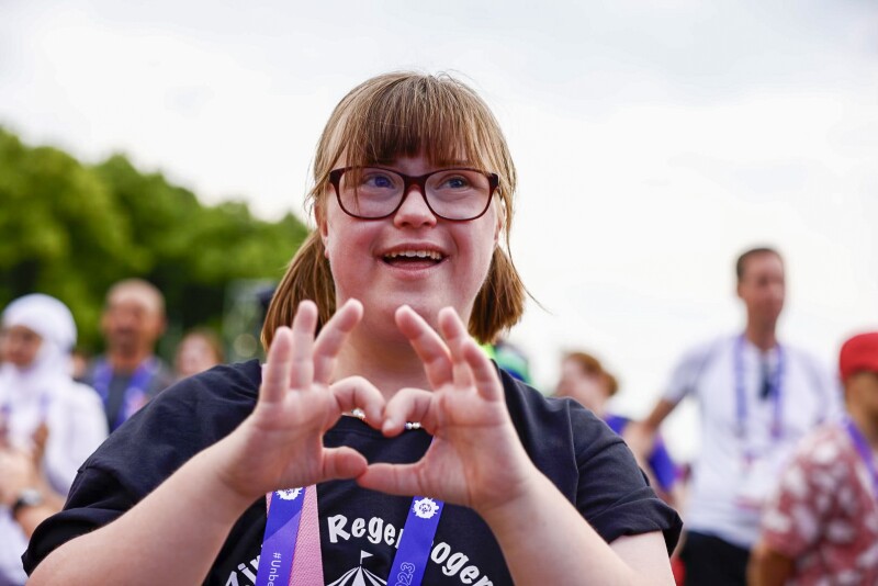 The photo shows a girl in a crowd. She is forming a heart with her hands to the camera and is laughing.