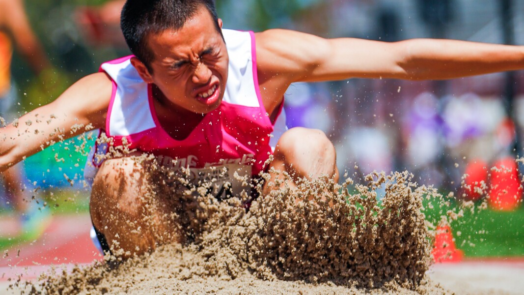 Das Bild zeigt einen Athleten beim Weitsprung. Es spritzt viel Sand hoch und der Athlet strengt sich an.
