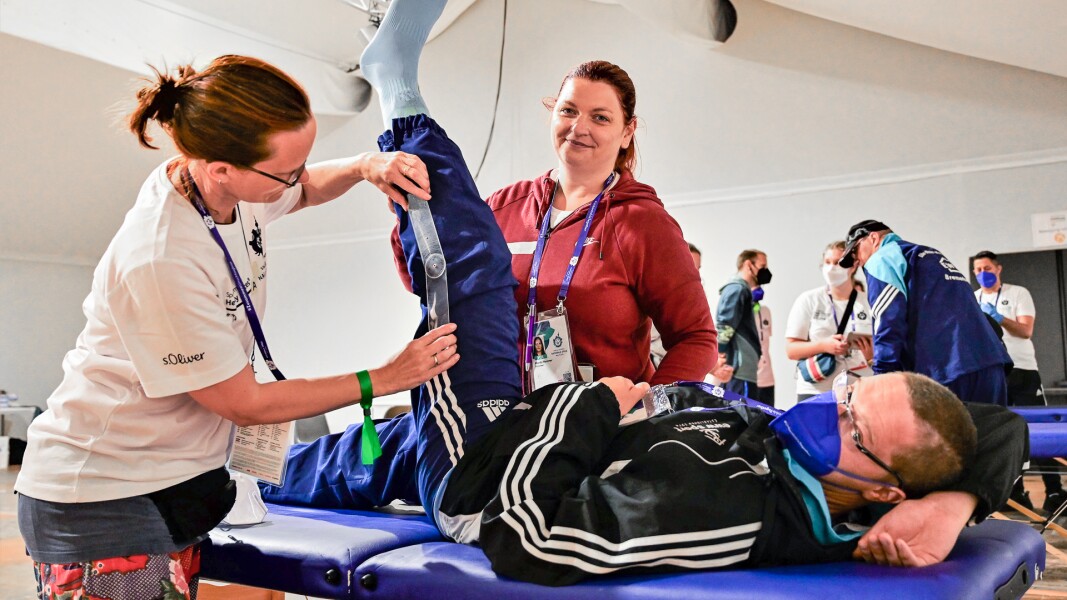 Auf dem Foto sind drei Menschen zu sehen. Ein Athlet liegt während einer Healthy Athletes Untersuchung auf einer Liege während die Beweglichkeit seines Beins durch eine Physiotherapeutin getestet wird. Daneben steht außerdem seine Betreuerin.