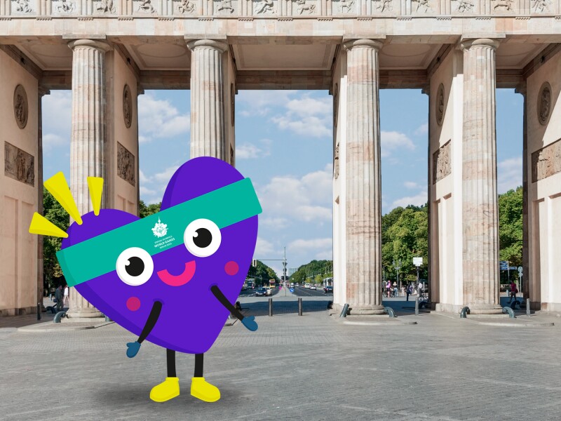 The photo shows the Special Olympics World Games Berlin 2023 mascot in front of the Brandenburg Tor. It has its arms outstretched and joyful eyes.