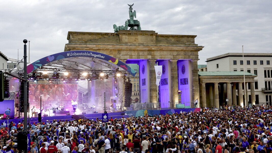 The photo shows the athletes party. A stage is set up in front of the Brandenburg Gate and a large crowd is gathered around it.