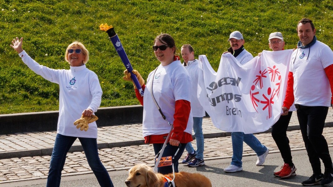 The photo shows a scene of the Bremen Torch Relay on the symbolic way to the Special Olympics National Games Berlin 2022. A total of 6 people and a companion dog can be seen. The happy smiling people are carrying a Special Olympics flag and one athlete is carrying the torch.