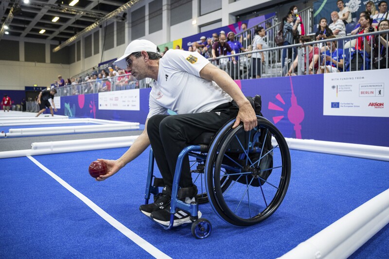 EN: German athlete in black pants and white jersey in a wheelchair playing Bocce for this national team.