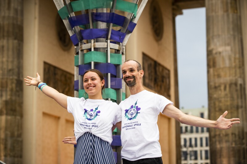 Auf dem Foto sieht man unsere beiden „Gesichter der Spiele": Lilly Binder und Robert Herberg. Sie posieren lächelnd und mit ausgebreiteten Armen vor der großen, frisch gelöschten Fackel vorm Brandenburger Tor im Rahmen der Abschlussfeier.