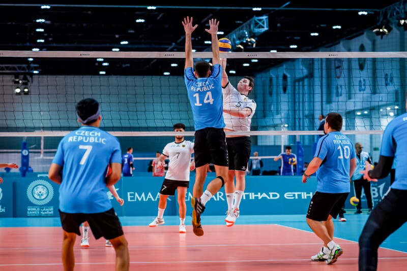 The photo shows two teams playing volleyball. Two players from the two teams are at the net. One is trying to pass the ball over the net, the other has his arms up and is trying to defend the ball.