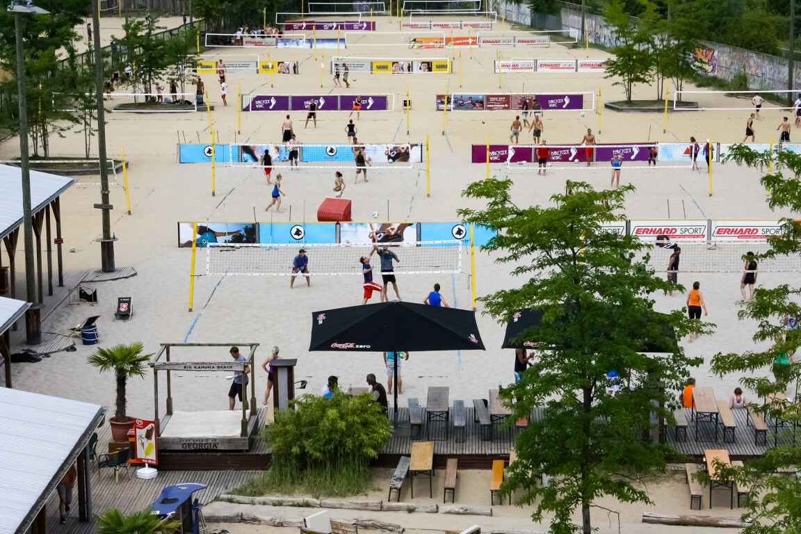 The photo shows a seating area with benches in the foreground and several beach volleyball courts where some people are playing beach volleyball.