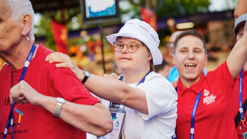 Auf dem Foto sind drei Menschen zu sehen, die an der Eröffnungsfeier der Nationalen Spiele in Berlin teilnehmen und Spaß haben. In der Szene nehmen sie an einer Tanz-Polonaise teil. 