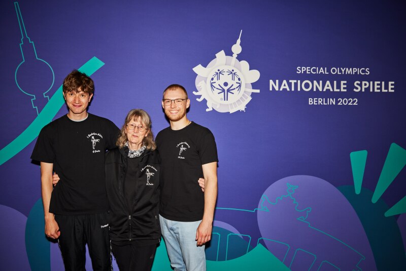 Auf dem Foto sieht man das Unified Team mit ihrer Trainerin von links nach rechts: Athlet Thorben von Kneten, Trainerin Gabriele Gramsch und Partner Lukas Ehresmann. Sie stehen Arm in Arm vor einer blauen Wand auf der das Special Olympics Nationale Spiele Berlin 2022 Logo ist.
