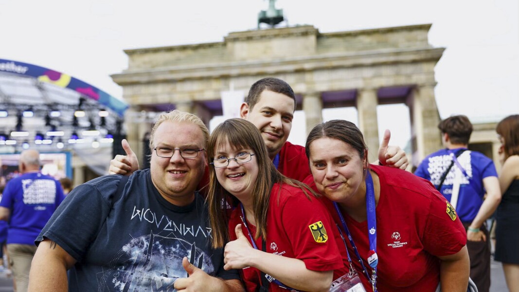 The photo shows four people in front of the Brandenburg Gate, including team members of the German delegation. They are laughing and giving a thumbs up to the camera.