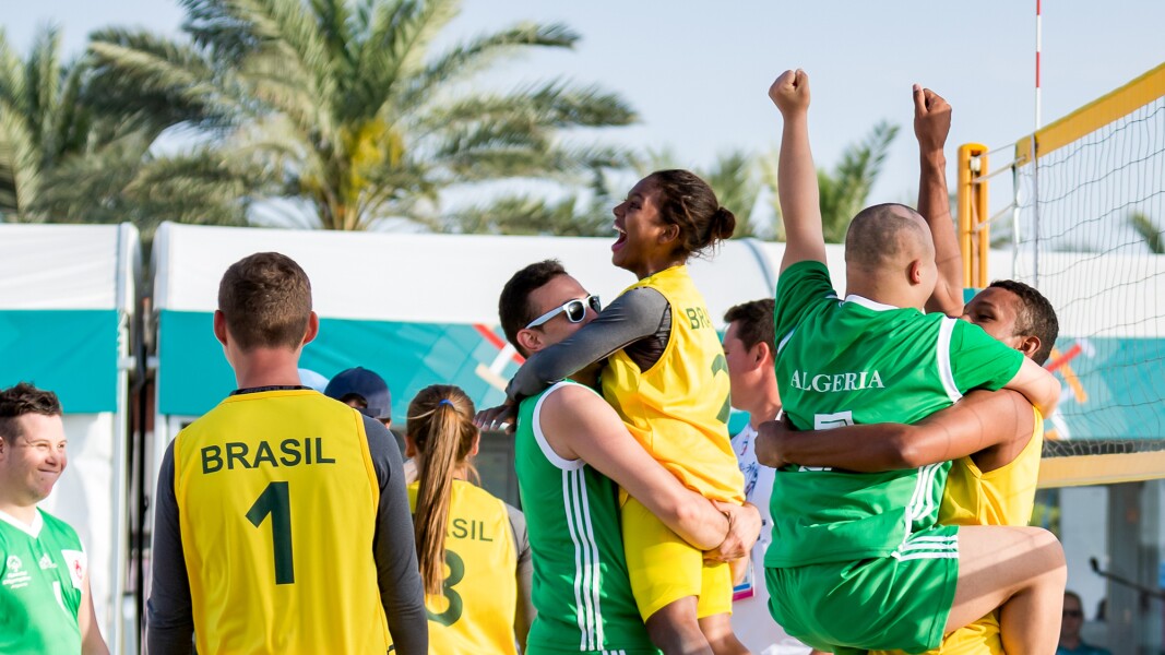 Auf dem Foto sind zwei freudige Mannschaften beim Beachvolleyball zu sehen, die sich gegenseitig in den Armen liegen und das Spiel feiern.