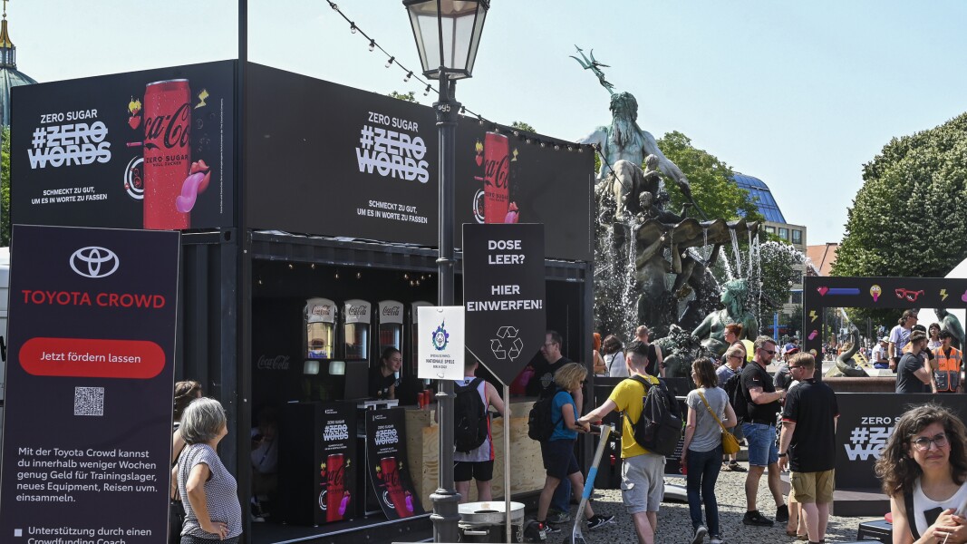 Das Foto zeigt verschiedene Sponsorenstände im Rahmen des Special Olympics Festivals. Im Vordergrund sieht man einen Aufsteller von Toyota, dahinter steht ein Truck der die CocaCola zero Sugar bewirbt. Es laufen viele Menschen umher.