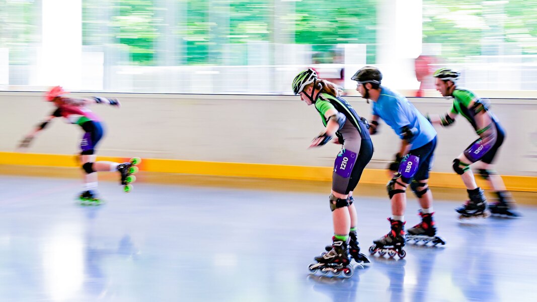 Auf dem Foto sieht man vier Roller Skater während ihrem Wettbewerb. Eine Athletin liegt in Führung, drei weitere Athleten liegen hinter ihr in etwa gleichauf.