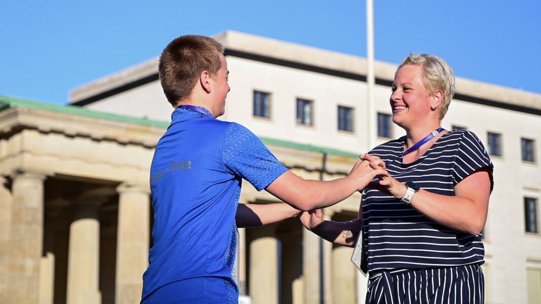 On the photo an athlete and a woman are dancing together. They stand in front of each other , hold themselves on their hands and smile. In the background you see a little piece of the Brandenburg Gate.