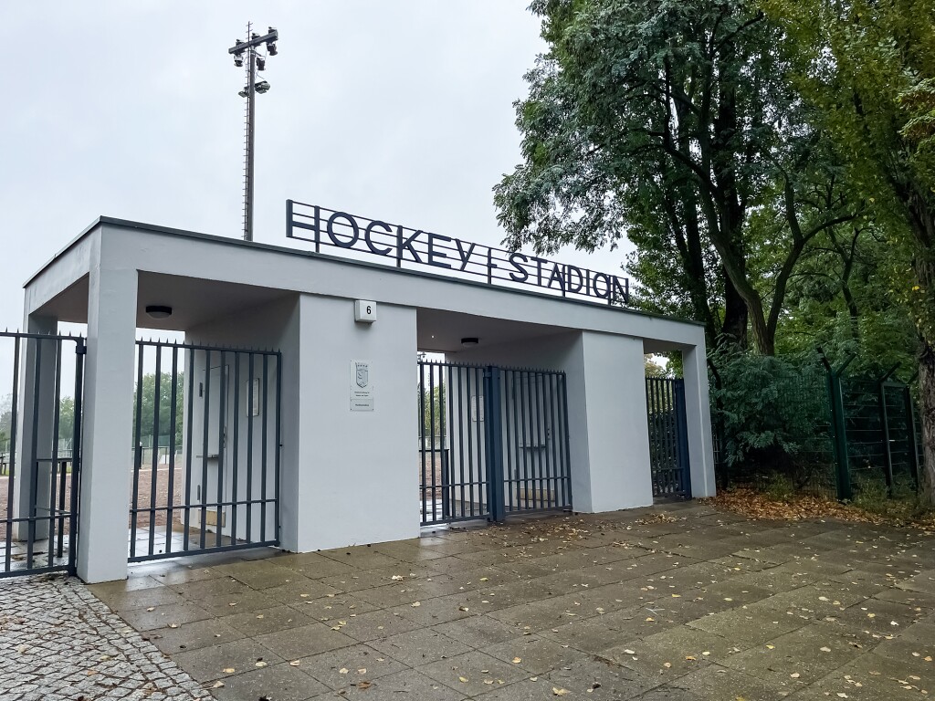 The photo shows an outdoor entrance area, with white columns and lattice gates. In front of it is a paved square.