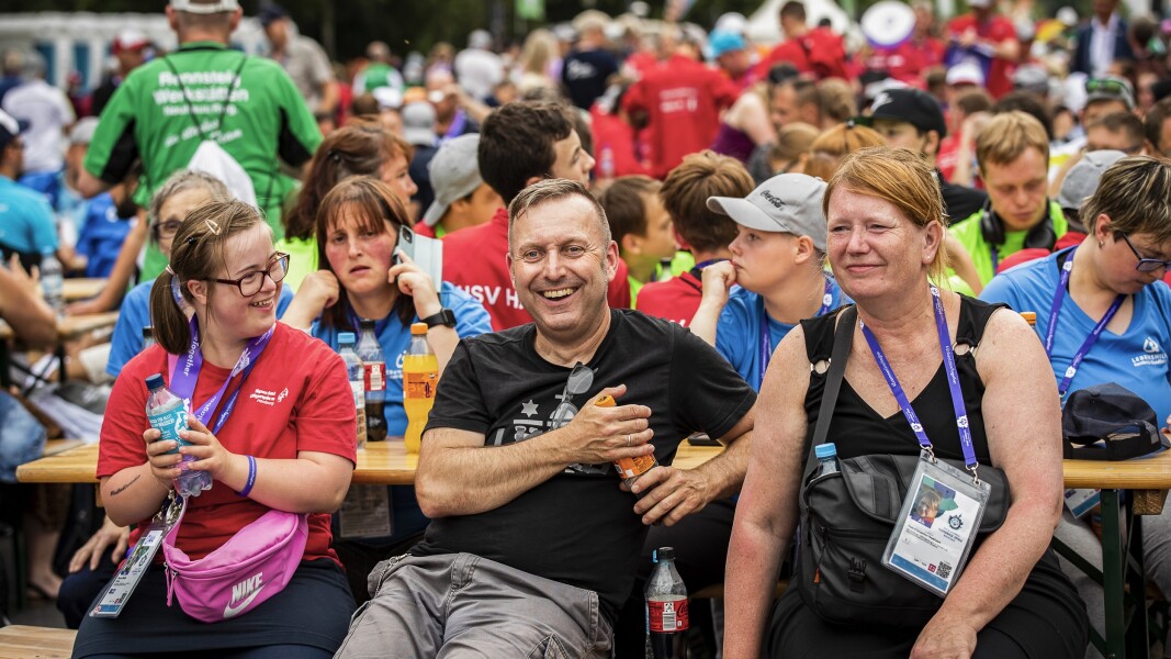 Auf dem Foto sieht man eine Szene der Abschlussfeier der Nationalen Spiele Berlin 2022 direkt am berühmten Brandenburger Tor. Im Vordergrund sitzen eine Athletin und ihre Eltern, alle drei lächeln und haben sichtlich Spaß.