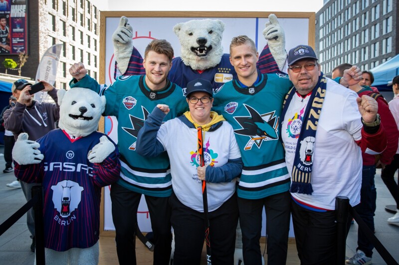 Auf dem Foto sind das Maskottchen der Eisbären Berlin, zwei Spieler und zwei Repräsentant*innen von Special Olympics zu sehen. Alle jubelns freudig in Richtung Kamera. 