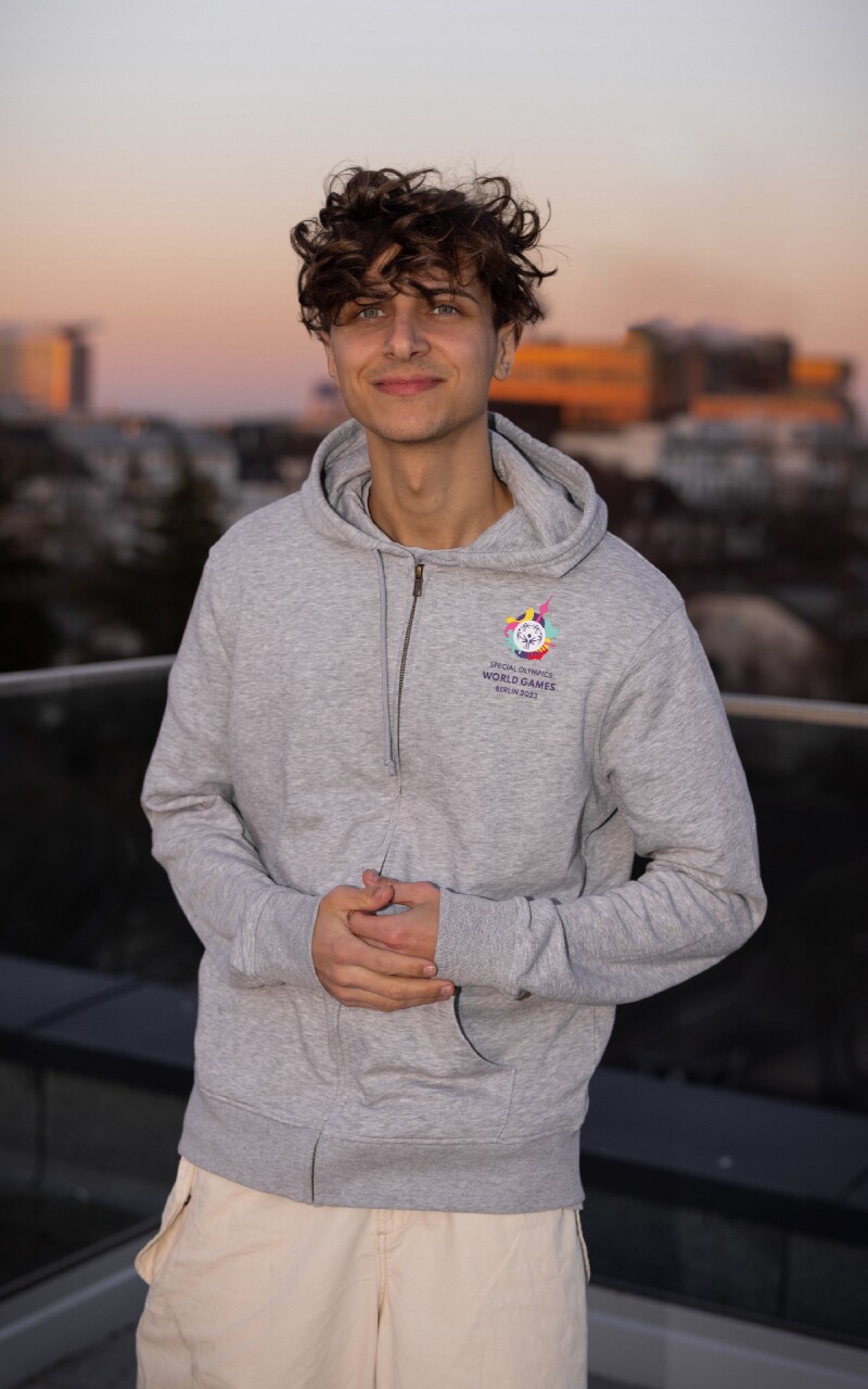 Das Foto zeigt den deutschen Popsänger Lukas Rieger. Er trägt einen grauen Weltspiele-Hoodie und lächelt in Richtung der Kamera.