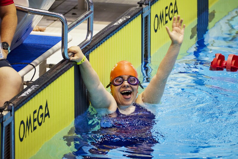 The photo shows a Spanish swimmer at the finish of the pool. She is holding onto the starting block with one hand while cheering with the other. She looks very happy doing this!