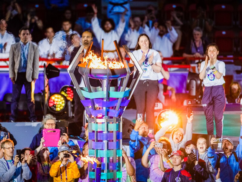 The photo shows a scene of the Opening Ceremony of the National Games in Berlin. In the center is the big torch flame and around it are some people taking photos of it while cheering.