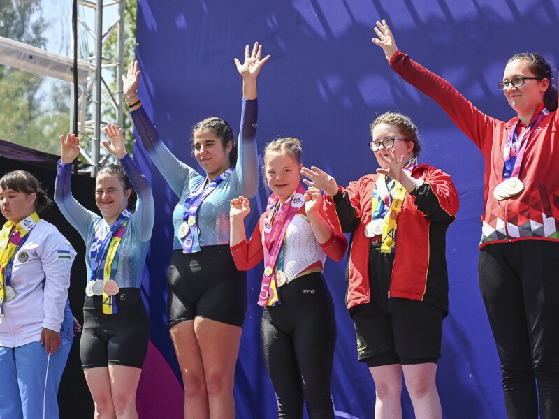 Auf dem Bild stehen 6 Athetinnen auf dem Podium und freuen sich auf über die gewonnen Medaillen. Alle jubeln vor Freude mit gestreckten Armen nach oben in Richtung des Publikums.