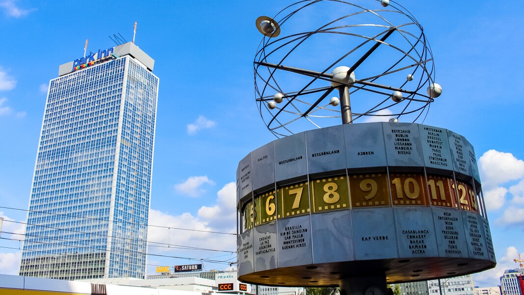 Das Foto zeigt die Weltzeituhr am Alexanderplatz in Berlin im Vordergrund. Im Hintergrund ist eine Tram und ein Hochhaus zu sehen.