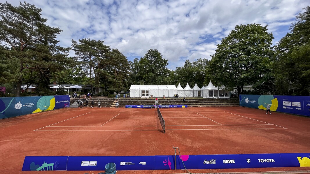 Das Foto zeigt den Hauptplatz (Center Court) der Tennisanlage. Es ist ein schöner Sandplatz umgeben von blauen Seitenaufstellern mit dem Logo der Special Olympics. Die Sonne scheint ein bisschen.