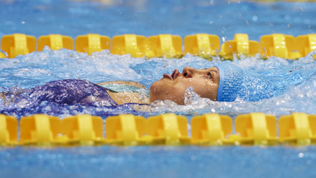 Eine Schwimmerin schwimmt in Rückenlage auf der Distanz von 50 Metern. Sie trägt eine blaue Badekappe. Quer auf dem Bild sind die Schwimmbahnen durch gelbe Schwimmkörper abgegrenzt zu sehen.
