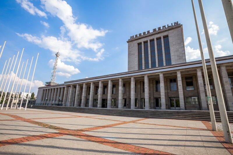 Das Foto zeigt einen gepflasterten Vorplatz und ein Gebäude mit Säulen auf dem Messe Berlin steht.