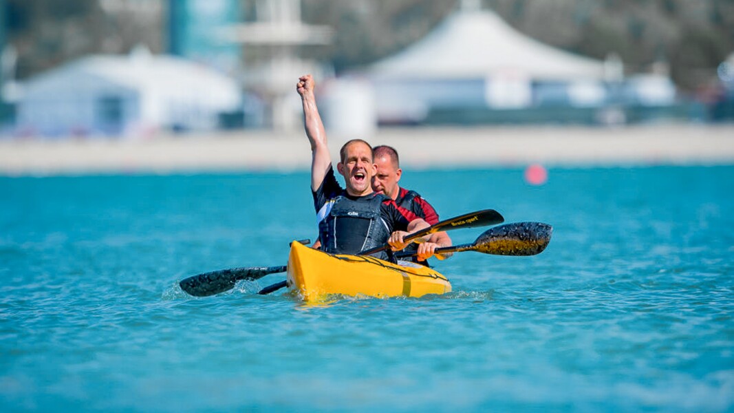 On the photo, there are two athletes in a kayak. The one up front is raising his fist in joy.