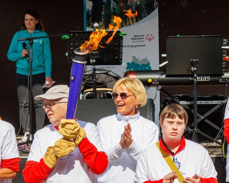 Das Foto zeigt eine Gruppe Menschen, die am Bremer Fackellauf auf dem symbolischen Weg zu den Nationalen Spielen Berlin 2022 teilnehmen. Ein Mann trägt die Fackel und alle klatschen in die Hände. Hinter der Gruppe ist eine Bühne aufgebaut.